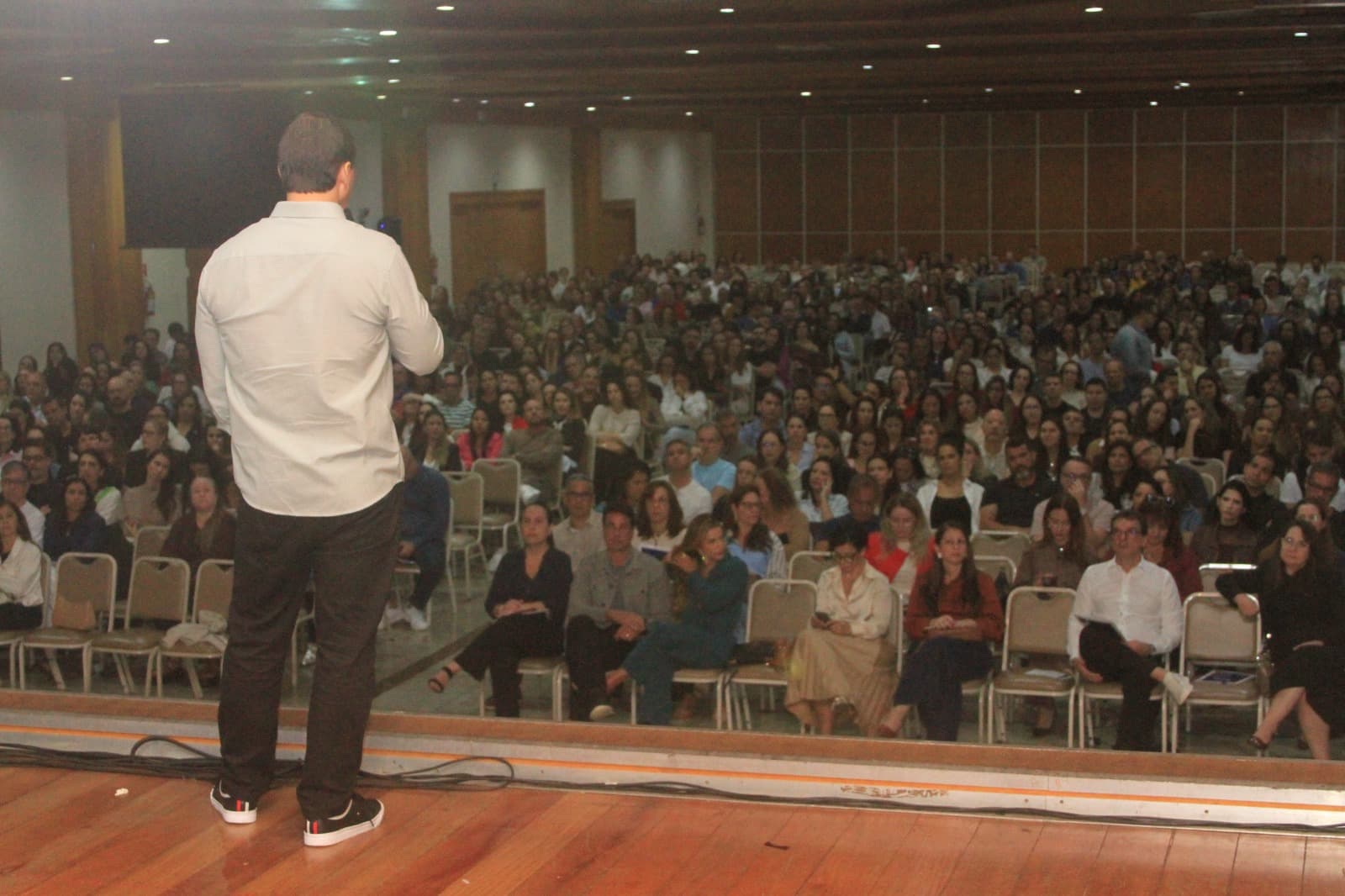Eduardo Pinheiro no palco de um auditório lotado, de costas para a câmera, falando para a plateia